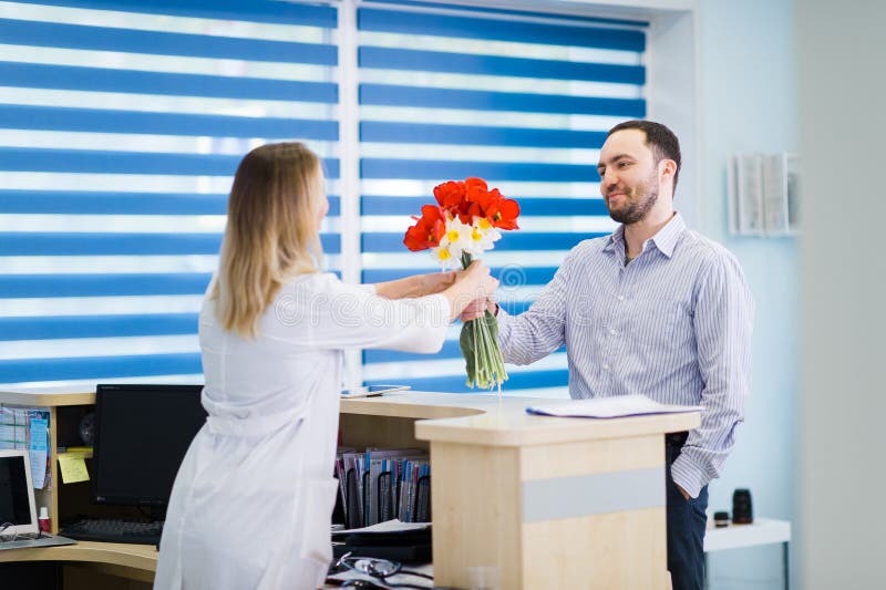 Una chica joven hermosa en una capa blanca se está colocando cerca del escritorio en la oficina y está tomando el boquet de flore fotos de archivo libres de regalías Una chica joven hermosa en una capa blanca se está colocando cerca del escritorio en la oficina y está tomando el boquet de flore fotos de archivo libres de regalías