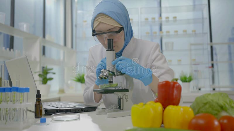 Una científica en una bufanda musulmana estudia verduras en un microscopio