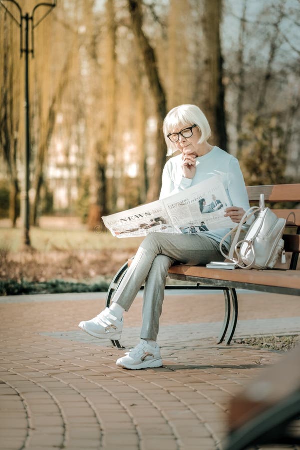 Una donna allegra e sovraordinata con occhiali che legge un giornale sulla panchina fotografia stock libera da diritti Una donna allegra e sovraordinata con occhiali che legge un giornale sulla panchina fotografia stock libera da diritti