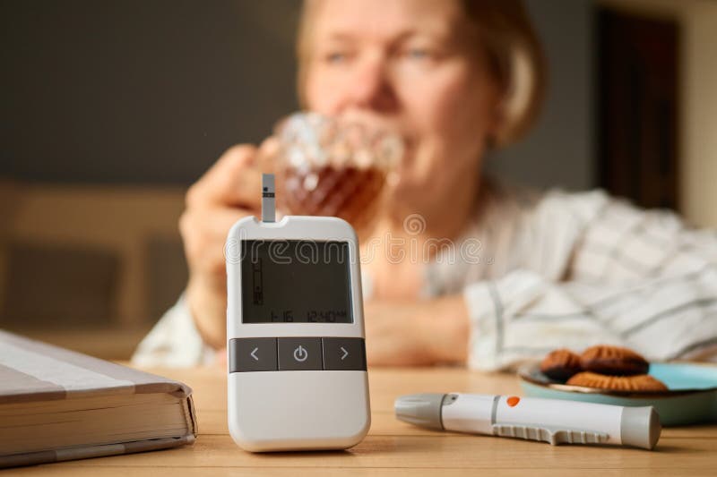 Una donna anziana siede silenziosamente mentre sorseggia il tè che controlla il suo zucchero nel sangue con un congegno messo sul immagini stock