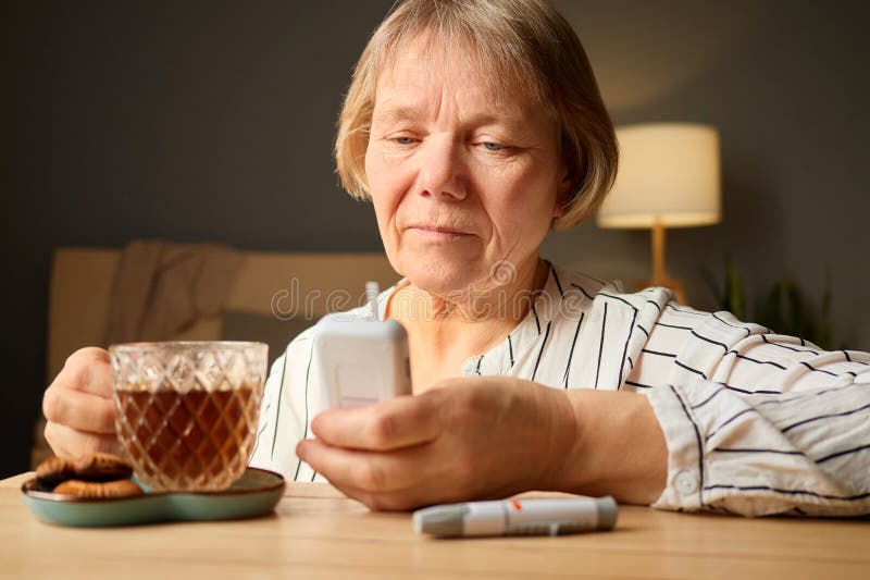 Una donna anziana siede in un tavolo di legno sorseggiando il tè da un bicchiere mentre controlla il telefono. I biscotti sono col immagini stock libere da diritti