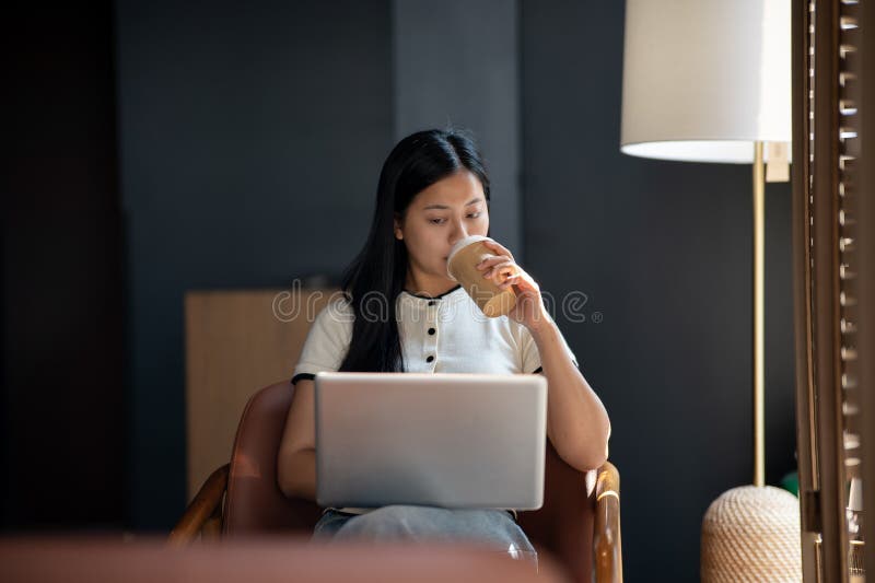 Una donna asiatica positiva lavora in remoto da una caffetteria leggendo attentamente i documenti sul suo portatile fotografia stock libera da diritti