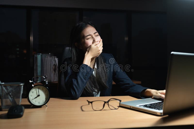 Una donna d'affari asiatica siede seduta sbadigliando con una mano coperta di bocca, il laptop, sulla scrivania, mentre la giovan fotografia stock