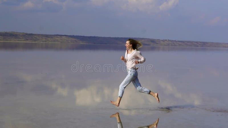 Una donna dai capelli lunghi e felici a casual corre sulla riva sabbiosa del lago con riflesso specchio