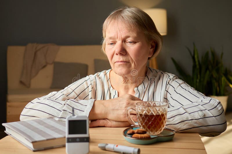 Una donna di età superiore si siede a un tavolo di legno osservando intensamente un glucometro. A lei piace una tazza di tè assiem. A lei piace una tazza di tè insieme ai biscotti che creano un'atmosfera calda nel suo spazio vitale.