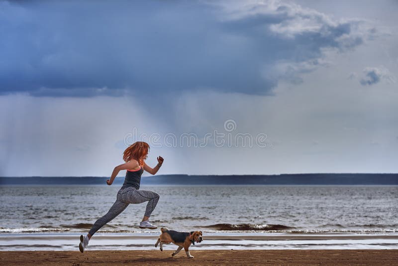 Una donna di mezza età dai capelli rossi nei funzionamenti degli abiti sportivi lungo la riva sabbiosa di grande fiume con il suo fotografia stock Una donna di mezza età dai capelli rossi nei funzionamenti degli abiti sportivi lungo la riva sabbiosa di grande fiume con il suo fotografia stock