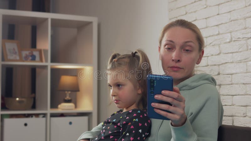 Una donna e una bambina sono sedute sul divano a casa una donna sta guardando lo schermo dello smartphone.