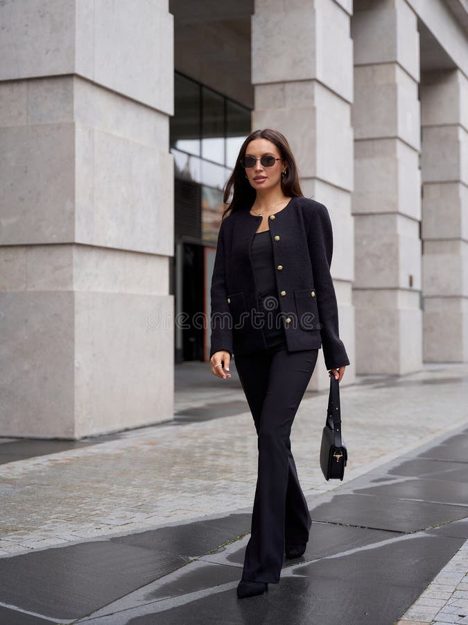 Una donna elegante in completo nero, composta da una giacca e pantaloni, cammina fiduciosamente su pavimentazione di pietra. Lei h fotografie stock