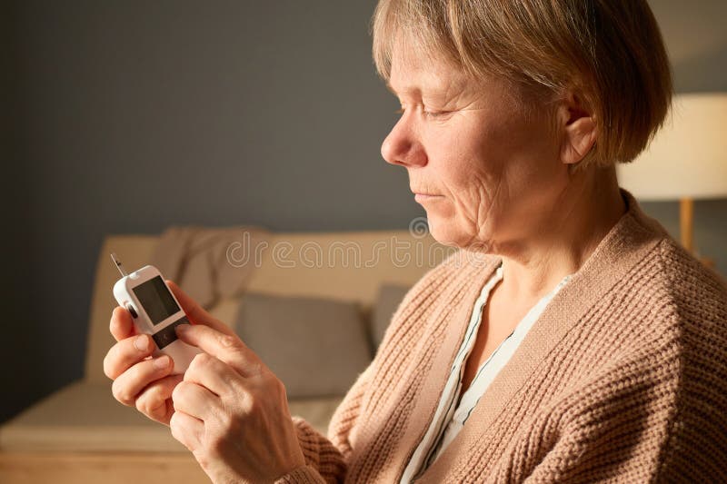 Una donna siede in una stanza confortevole, concentrata sul monitoraggio dei livelli di glucosio con un dispositivo. Sembra premur fotografia stock