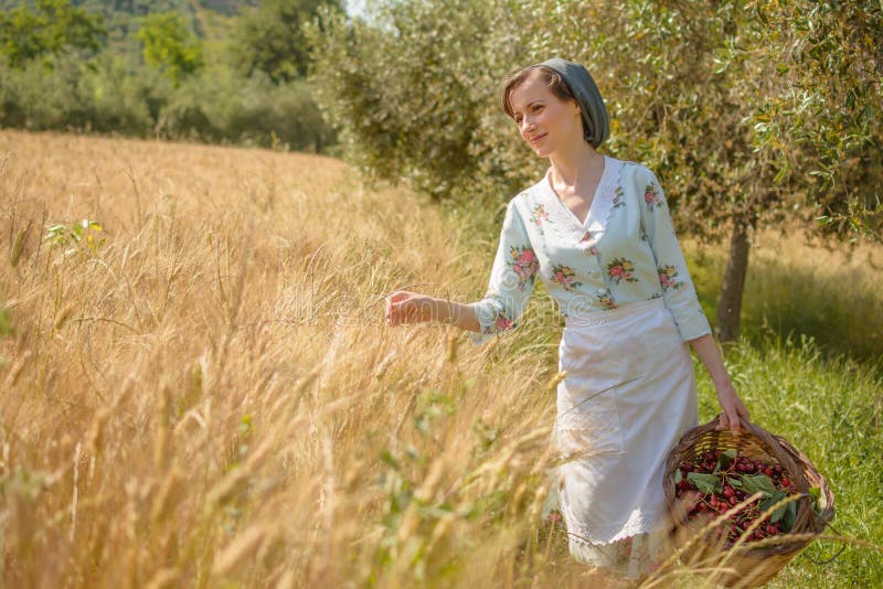 Una donna vestita da quarant'anni cammina nella campagna italiana, accanto a un campo di grano, portando un cesto di ciliegie