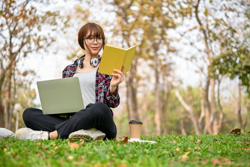 Focused and attractive young Asian female college student in casual clothes sits on the grass in the greenery park,reads a book,and uses her laptop to manage her school work. Focused and attractive young Asian female college student in casual clothes sits on the grass in the greenery park,reads a book,and uses her laptop to manage her school work