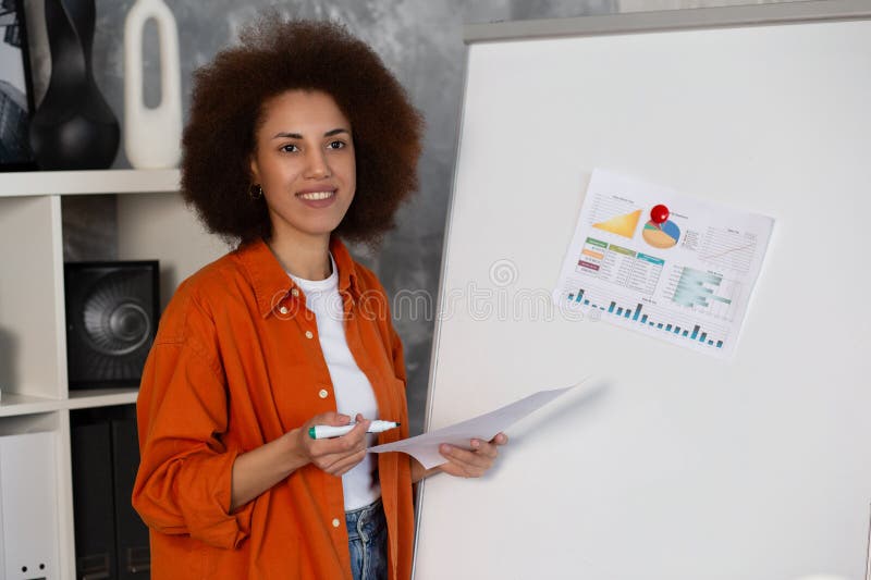 Smart successful business african american woman, top manager, stands in the modern office near the marker board, analyze financial charts, profits, strategy. Smart successful business african american woman, top manager, stands in the modern office near the marker board, analyze financial charts, profits, strategy