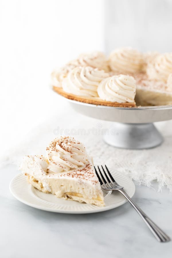 Una fetta di torta alla crema di banane con la torta piena dietro contro una finestra di sole brillante. fotografia stock libera da diritti