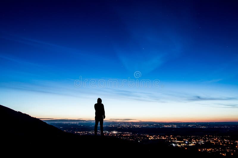 Una figura encapuchada solitaria en una colina silueteada momentos antes de la salida del sol que mira hacia fuera en ciudad se e imágenes de archivo libres de regalías Una figura encapuchada solitaria en una colina silueteada momentos antes de la salida del sol que mira hacia fuera en ciudad se e imágenes de archivo libres de regalías