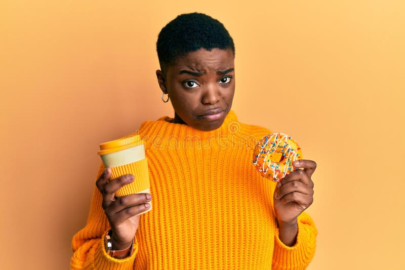 Una giovane donna africana americana che mangia ciambella e beve toglie il caffè scettico e nervosa accigliato a causa del problem fotografie stock