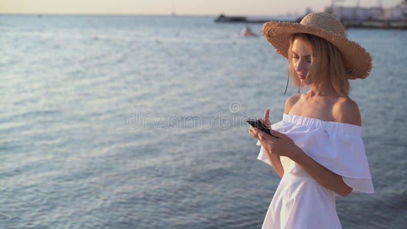 Una giovane donna caucasica cammina per il mare o l'oceano la mattina presto all'alba tenendo in mano uno smartphone che le esce d