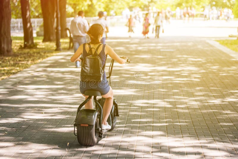 Una giovane donna che guida la bici elettrica del motorino di eco a emissione nulla in un parco della città fotografia stock Una giovane donna che guida la bici elettrica del motorino di eco a emissione nulla in un parco della città fotografia stock