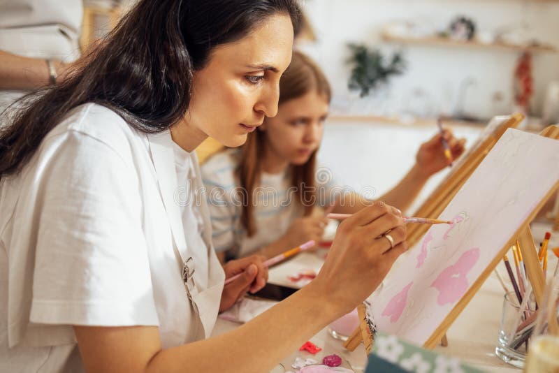 Una giovane donna concentrata in camicia bianca e grembiule dipingono un'immagine a un tavolo immagini stock