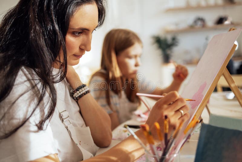 Una giovane donna concentrata con una maglietta bianca e un grembiule dipinge un'immagine a un tavolo e un alluce fotografia stock libera da diritti