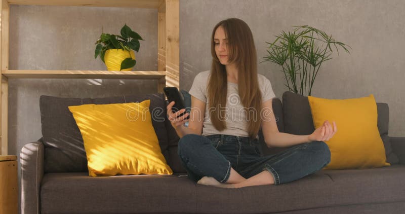 Una giovane donna suona la musica sul suo telefono e medita sul divano in soggiorno