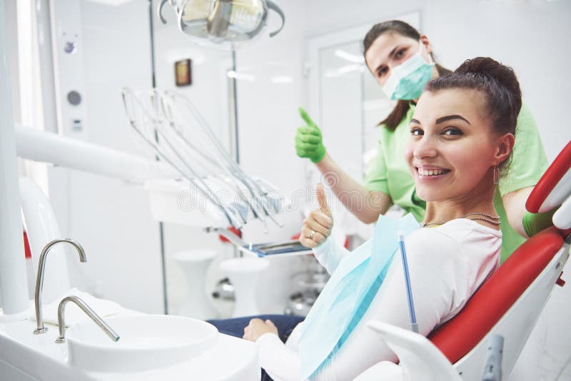 Una giovane donna è soddisfatta con il lavoro del dentista e sostiene il suo pollice immagini stock libere da diritti Una giovane donna è soddisfatta con il lavoro del dentista e sostiene il suo pollice immagini stock libere da diritti