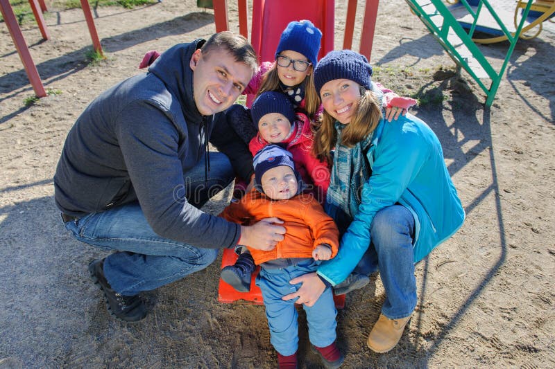 Una giovane famiglia di cinque sorridente felice al campo da giuoco del ` s dei bambini in parco fotografia stock Una giovane famiglia di cinque sorridente felice al campo da giuoco del ` s dei bambini in parco fotografia stock