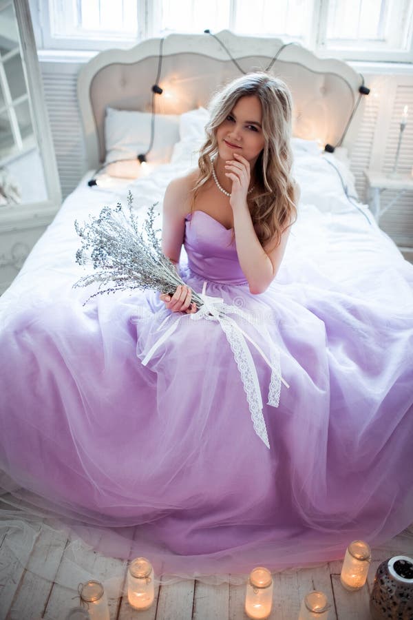 Una giovane sposa bionda in un vestito porpora magnifico che si siede sul letto Nelle candele della stanza Tenute della ragazza n fotografia stock Una giovane sposa bionda in un vestito porpora magnifico che si siede sul letto Nelle candele della stanza Tenute della ragazza n fotografia stock