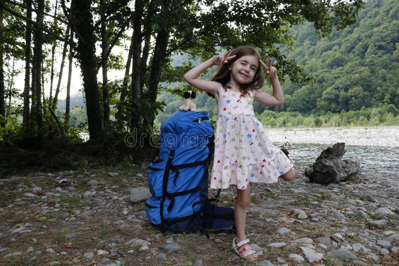 Una gran mochila para un adulto se para al lado de una niñita con el telón de fondo de árboles y un río en un día soleado. fotografía de archivo libre de regalías