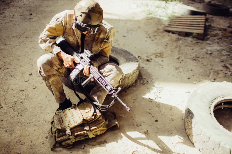 Una imagen del soldado que se sienta en la tierra y la mascarilla que lleva Él está sosteniendo el rifle negro en manos El hombre imagen de archivo libre de regalías Una imagen del soldado que se sienta en la tierra y la mascarilla que lleva Él está sosteniendo el rifle negro en manos El hombre imagen de archivo libre de regalías
