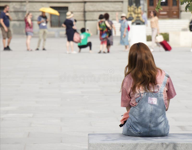 Una joven adolescente con jengibre en jeans sentada y esperando en el banco de la ciudad en la plaza, por detrás imagen de archivo Una joven adolescente con jengibre en jeans sentada y esperando en el banco de la ciudad en la plaza, por detrás imagen de archivo