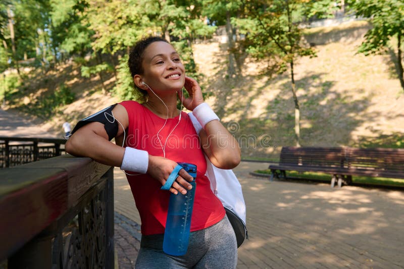 Una joven deportista en ropa activa sonríe hermosa toothy sonriendo sosteniendo una botella con agua y poniendo auriculares relajá fotografía de archivo libre de regalías
