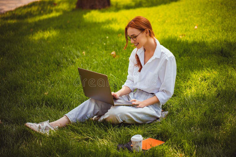 Una joven empresaria pelirroja sentada sobre el césped en el parque natural utiliza una laptop pc para trabajar fuera de la oficin fotos de archivo libres de regalías