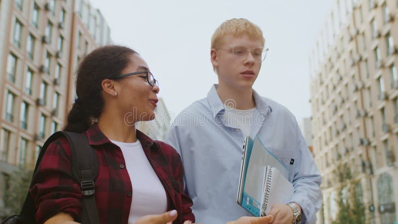 Una joven negra y un joven caucásico caminando por una calle de la ciudad y hablando con edificios de varios pisos como un almacen de metraje de vídeo