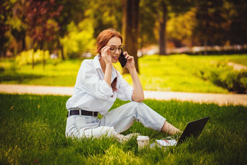 Una joven pelirroja trabaja con una laptop y un teléfono celular al aire libre mientras está sentada en la hierba. concepto independiente