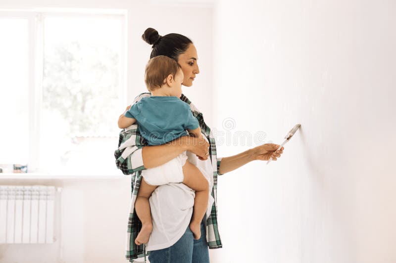 Una joven sostiene a un niño y pinta una pared. una encantadora madre con un niño pequeño está haciendo reparaciones en un apartam imagen de archivo libre de regalías