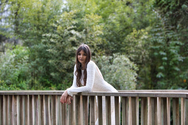 Una joven y hermosa morena vestida con un vestido de punto blanco se inclina en el pasamanos de madera de un puente fotografía de archivo libre de regalías
