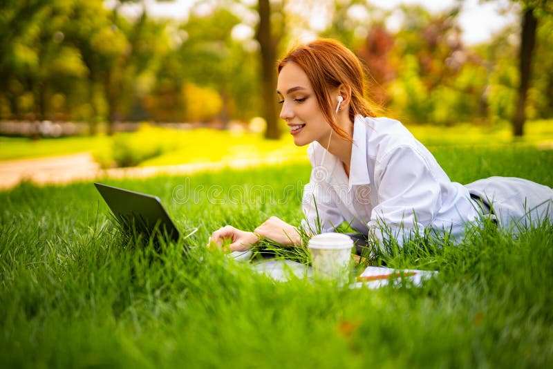 Una joven y hermosa pelirroja sonriente usa un ordenador portátil y audífonos mientras se sienta sobre el césped en el parque foto de archivo