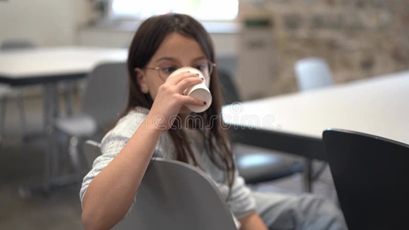 Una linda chica con gafas y un suéter bebiendo de una taza de papel se sienta en una silla frente a una mesa en una cafetería del almacen de metraje de vídeo
