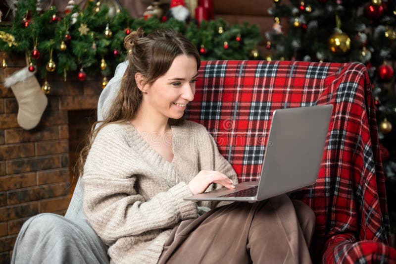 Una linda chica con un suéter está sentada en una silla roja cerca del árbol de navidad y trabajando en una laptop imágenes de archivo libres de regalías
