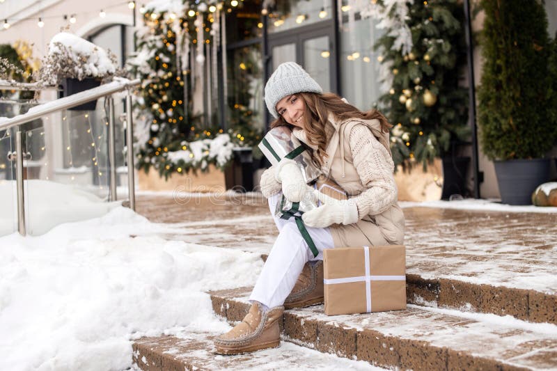 Una linda chica en mittens está sentada en los peldaños con nieve y sosteniendo regalos afuera en invierno foto de archivo libre de regalías