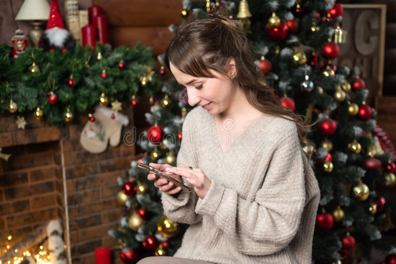 Una linda chica está sentada cerca del árbol de navidad, mirando el teléfono y leyendo un mensaje imagenes de archivo