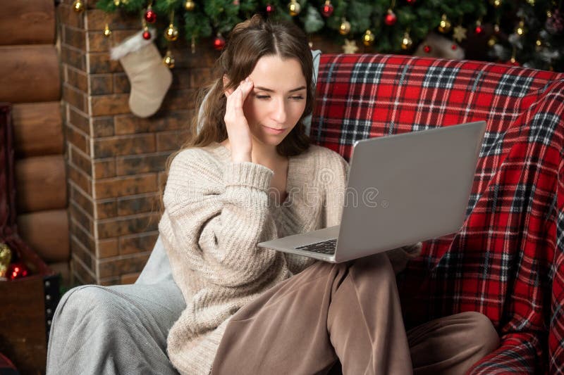 Una linda muchacha está sentada cerca del árbol de navidad experimentando una migraña y trabajando en una laptop foto de archivo libre de regalías