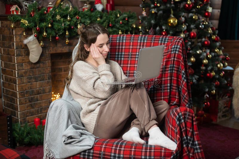 Una linda muchacha está sentada cerca del árbol de navidad experimentando una migraña y trabajando en una laptop fotografía de archivo libre de regalías