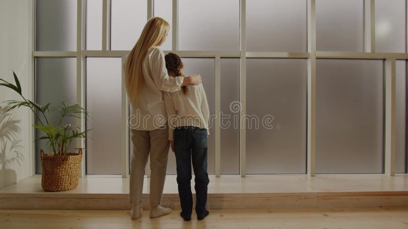 Una madre joven y su hija en crecimiento se paran en un abrazo y miran por una ventana grande. manifestación del amor materno almacen de metraje de vídeo