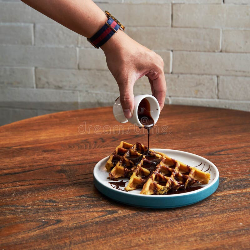 Una mano versa un ricco sciroppo di cioccolato su un piatto di waffe appena confezionate godendosi un dolce trattamento in un caff fotografie stock