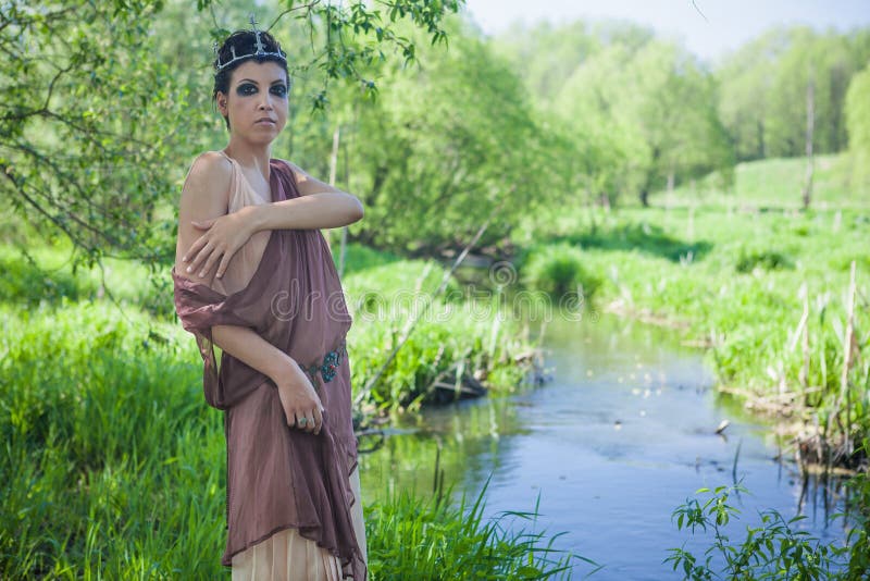 Una morenita fina en un vestido marrón con una corona en su cabeza miente en una rama de árbol cerca de una charca del bosque fotos de archivo Una morenita fina en un vestido marrón con una corona en su cabeza miente en una rama de árbol cerca de una charca del bosque fotos de archivo