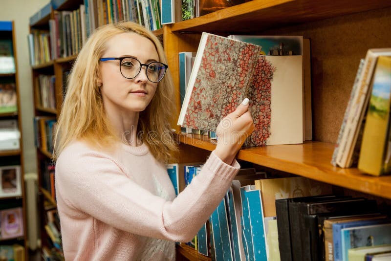 Una muchacha con los vidrios elige un libro en la biblioteca imagen de archivo Una muchacha con los vidrios elige un libro en la biblioteca imagen de archivo
