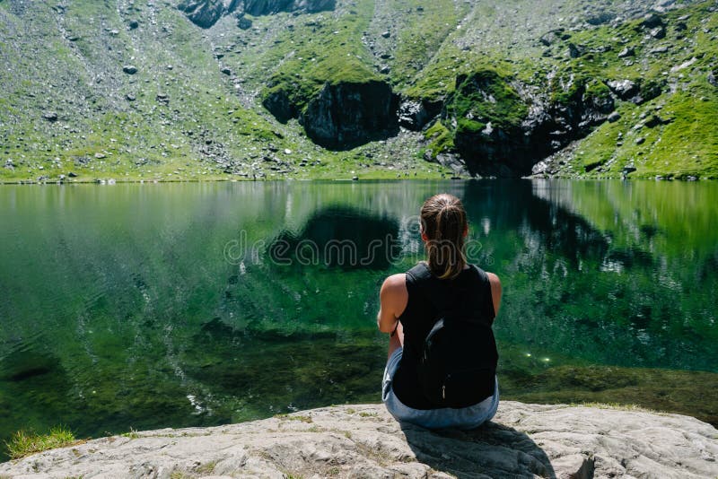 Una muchacha que se sienta en una roca, mirando una vista espectacular de la montaña que refleja en el lago