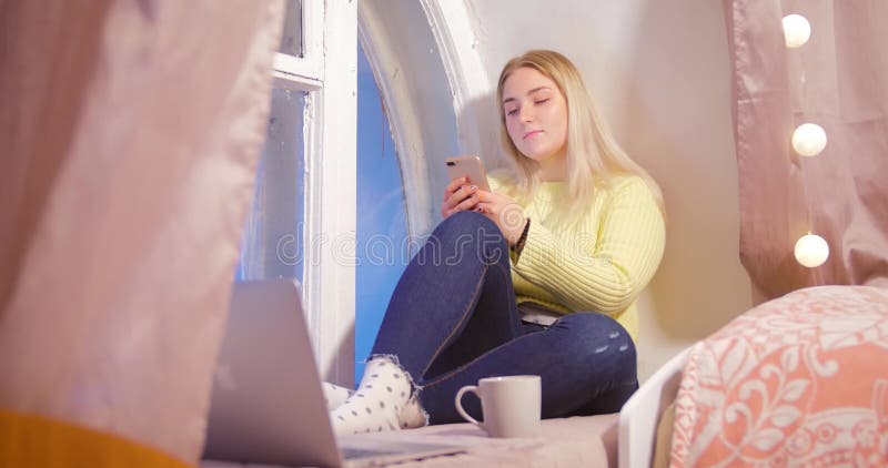 Una muchacha rubia en un suéter amarillo se está sentando con un teléfono cerca de la ventana en su sitio contra un fondo del ros almacen de video