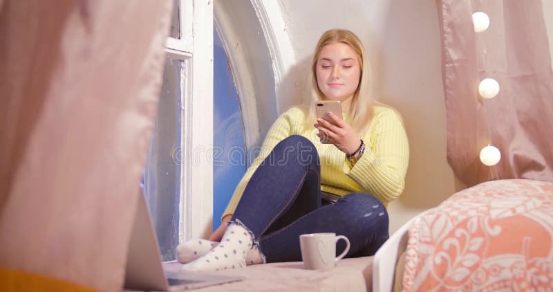 Una muchacha rubia en un suéter amarillo se está sentando con un teléfono en su sitio contra un fondo de cortinas rosadas y metrajes